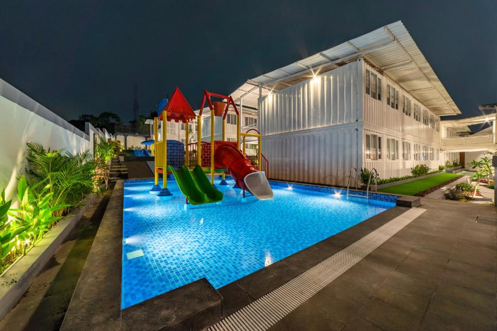 A bright blue swimming pool at night with a colorful children's playground slide in the middle, situated next to modern white container-style buildings with warm lighting.
