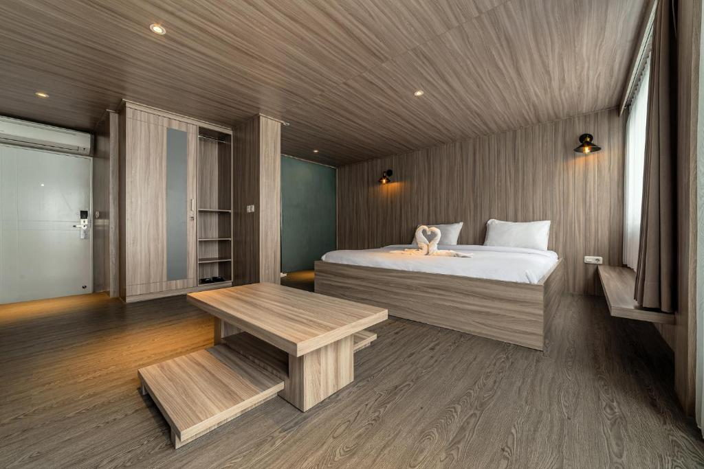 A modern hotel room interior with light-colored wood paneling on the walls, ceiling, and floor. It features a large bed with white linens, a wooden wardrobe, and a minimalist low wooden table.