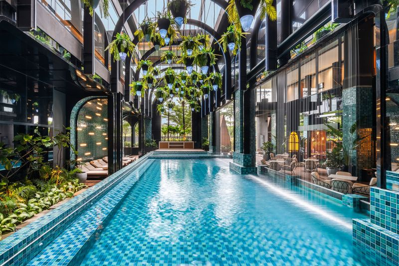 A long, luxurious semi-outdoor swimming pool with teal mosaic tiles. The area features high black industrial arches with many green plants hanging from the ceiling, surrounded by glass walls and lounge seating.