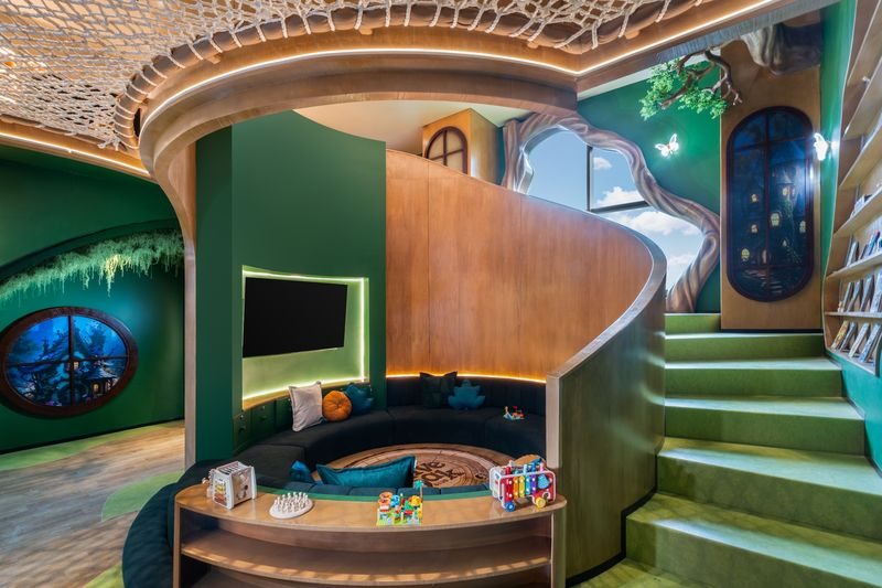 An indoor children's play area with a magical forest theme. It features curved wooden walls, a circular green seating pit with toys, green stairs, and a tree-like sculpture on the wall, resembling a Hobbit house.