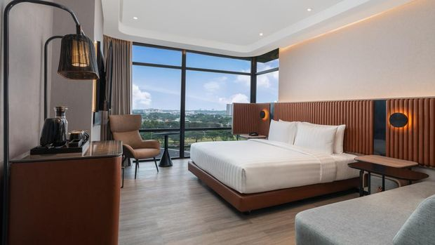 A modern hotel bedroom with a large bed, white linens, and a tall brown textured headboard. The room features a massive floor-to-ceiling window overlooking a city landscape under a blue sky, an armchair, and minimalist furniture.