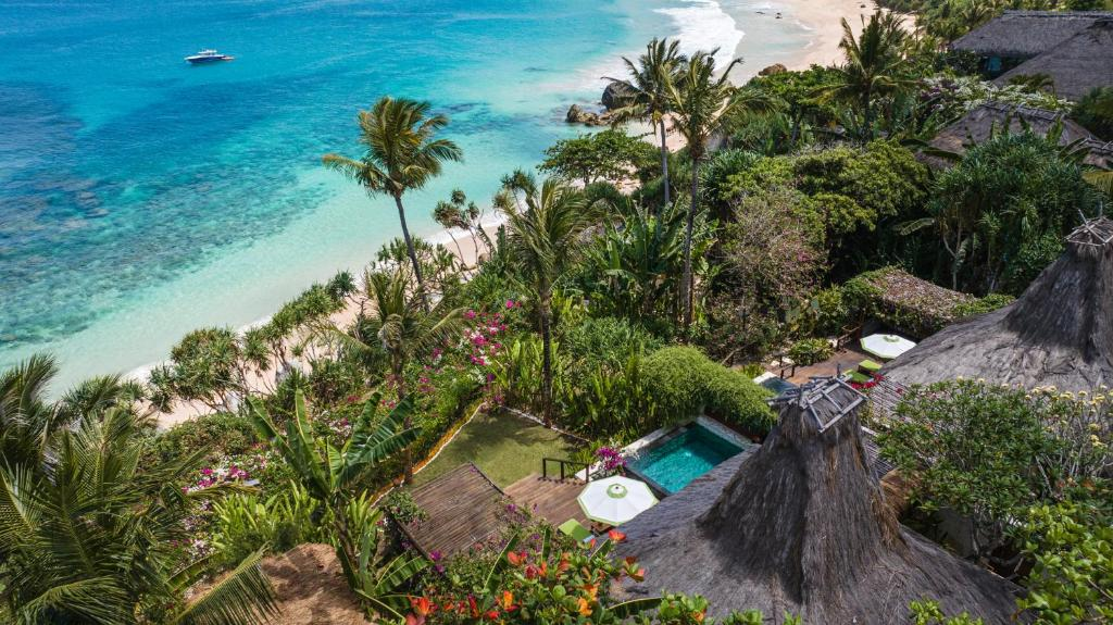 "A high-angle view of NIHI Sumba resort, featuring traditional thatched-roof villas with private infinity pools nestled among lush tropical greenery, overlooking a pristine white sand beach and crystal-clear turquoise ocean.