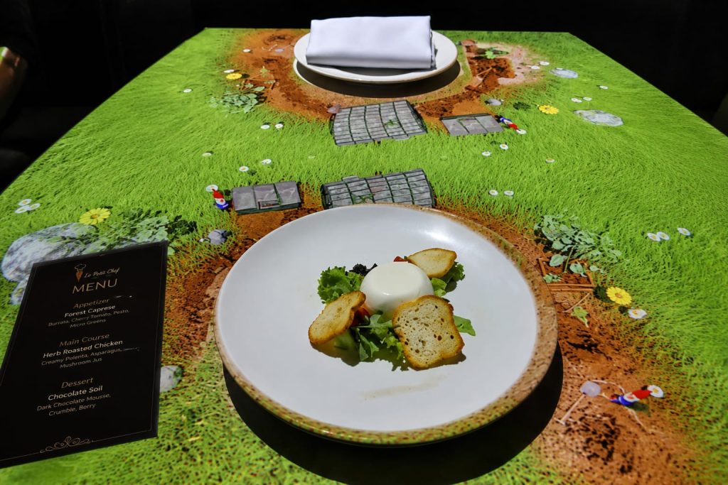 Piring berisi hidangan Forest Caprese di meja makan dengan proyeksi visual 3D bertema kebun dari pengalaman bersantap Le Petit Chef