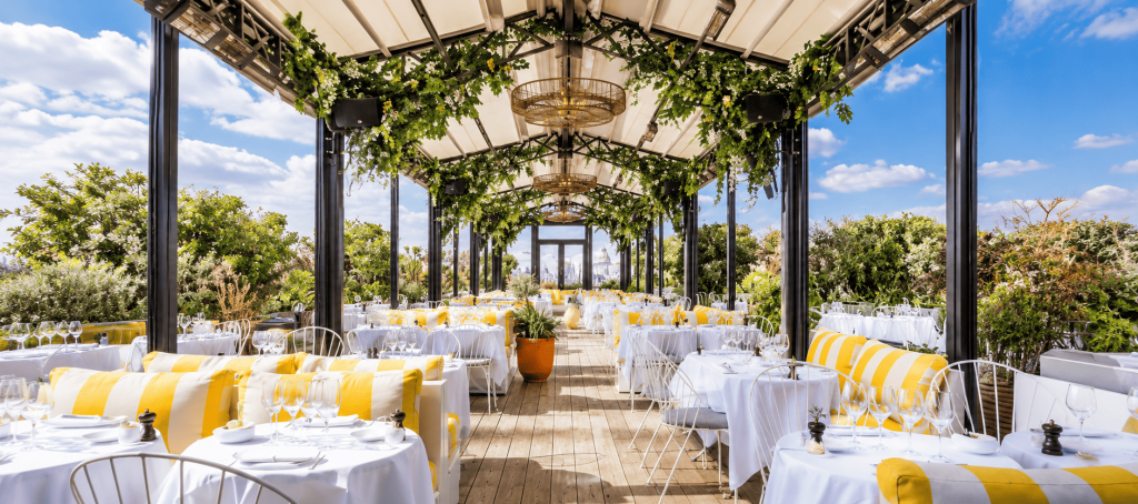 Area rooftop restaurant Perruche di Paris dengan meja makan berwarna putih, sofa kuning, tanaman hijau, dan panorama kota