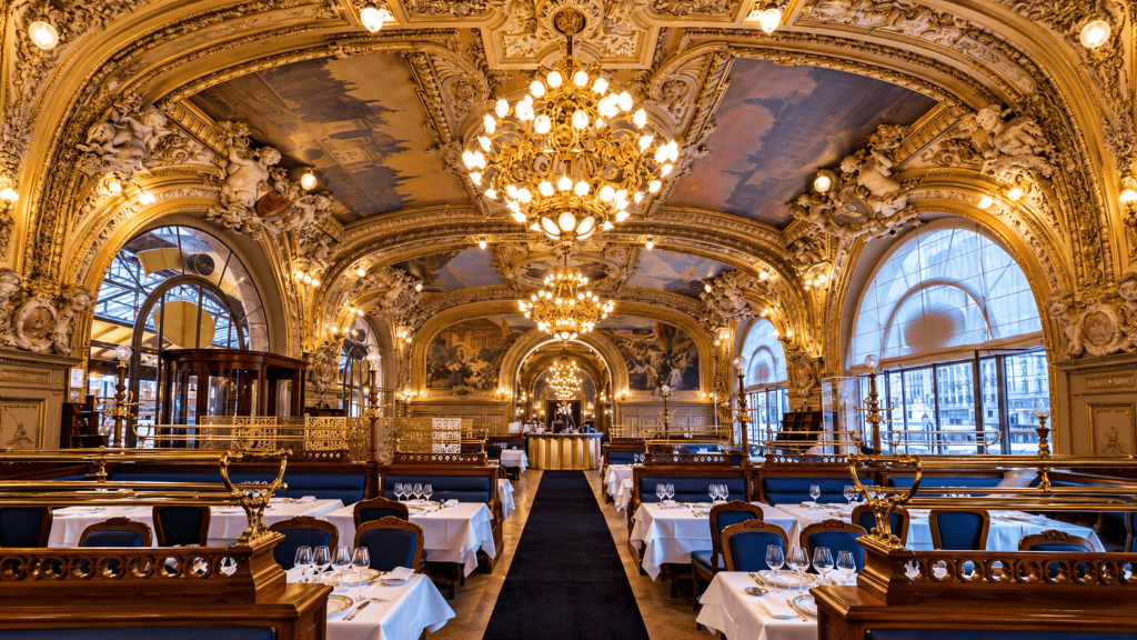 Interior Le Train Bleu di Paris dengan chandelier megah, ornamen emas, lukisan langit-langit, dan meja makan bergaya klasik