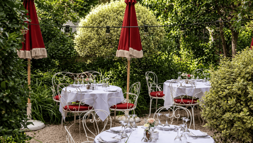 Area makan outdoor Hôtel Particulier Montmartre dengan meja ber taplak putih, kursi besi klasik, payung merah, dan taman hijau yang rimbun